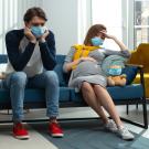 man and pregnant woman sitting on a couch wearing masks